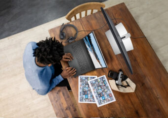 Overhead view of an African-American photographer working on photo editing on a Dell Precision 7000 series laptop, with a 27-inch Ultra HD 4K (P2721Q) monitor, photo proof pages and their DSLR camera.